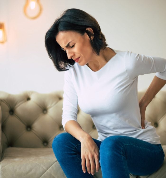 Axial pain. Close-up photo of a hurting woman, who is sitting on a couch and holding her lower back with her left hand.