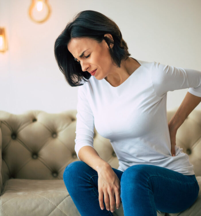 Axial pain. Close-up photo of a hurting woman, who is sitting on a couch and holding her lower back with her left hand.